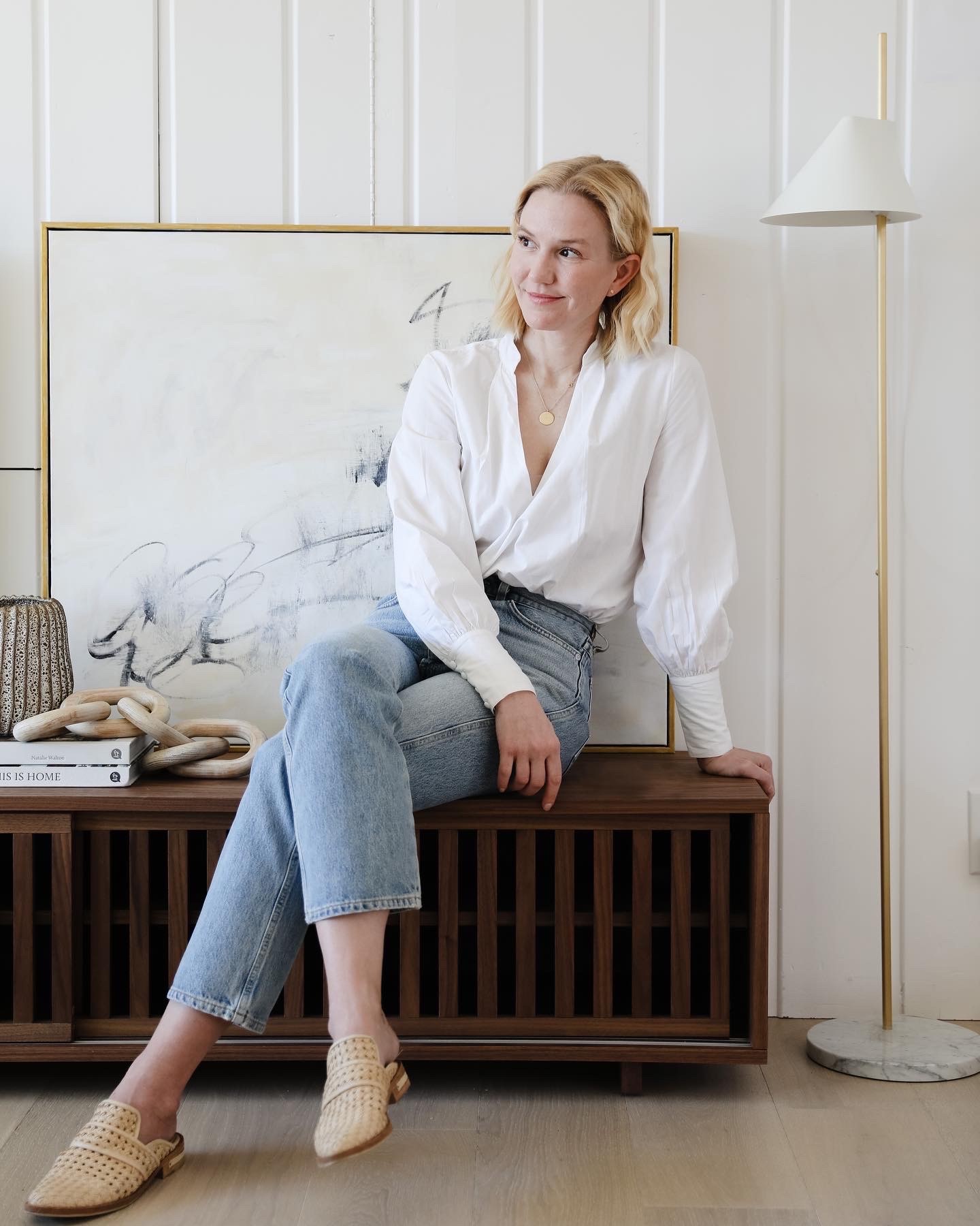 Woman white blouse and jeans in her living room, against a white panel wall.