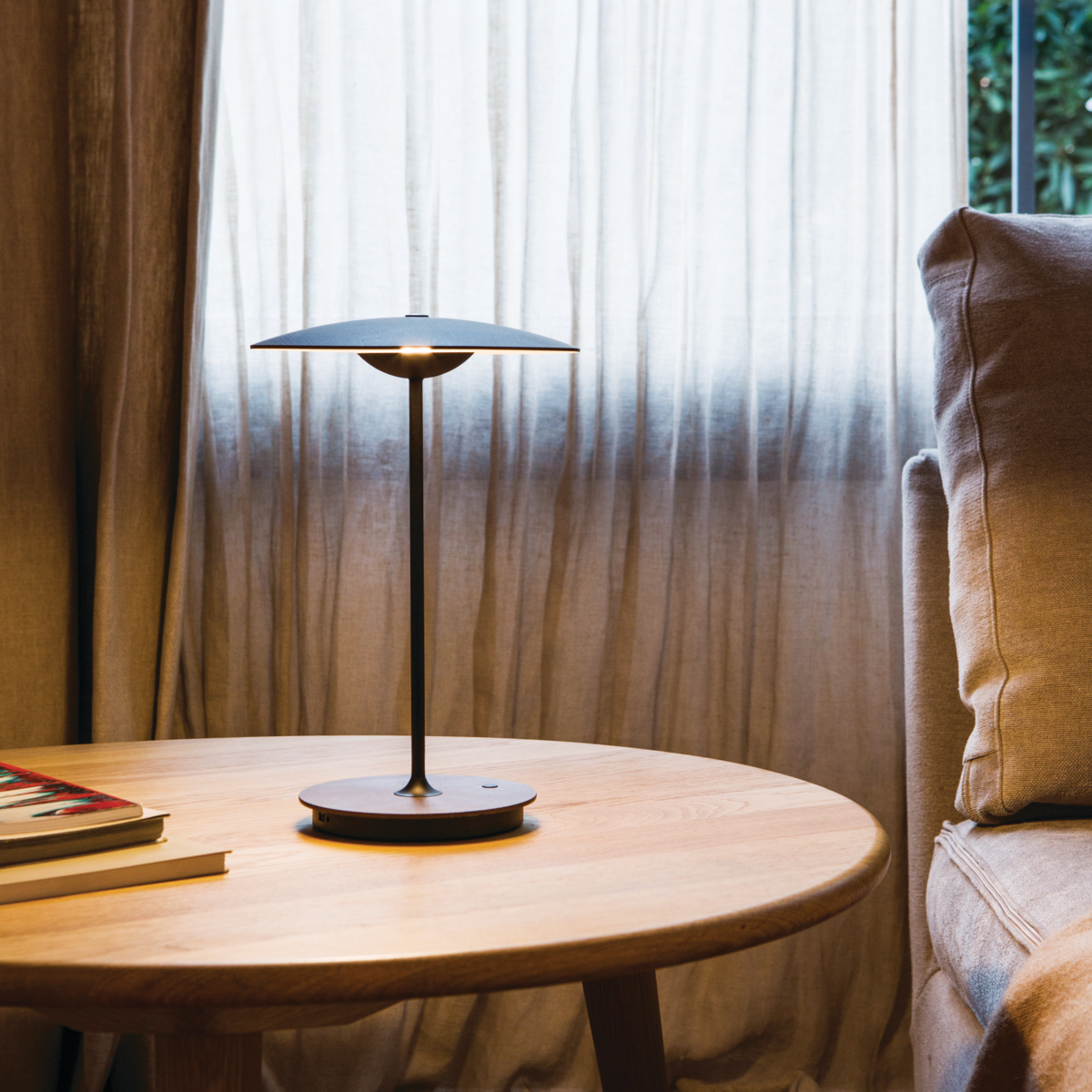 Small black table lamp on wood circular end table in front of a curtained window